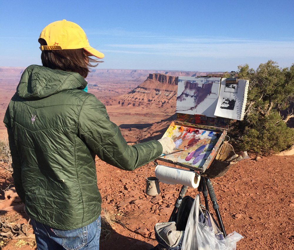 An artist painting on an easel outdoors with red cliffs off in the distance