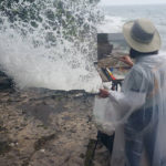 Woman artist painting outdoors right near crashing waves