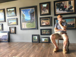 Artist sitting in his studio with framed paintings filling the wall