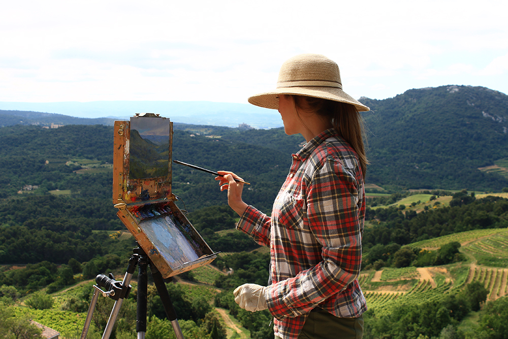 Female artist painting outside with a lush green landscape in the distance