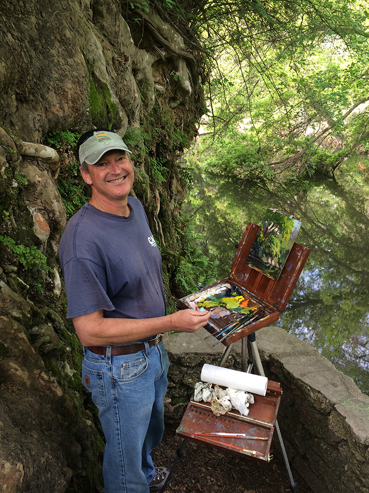 Male artist painting outside by a creek