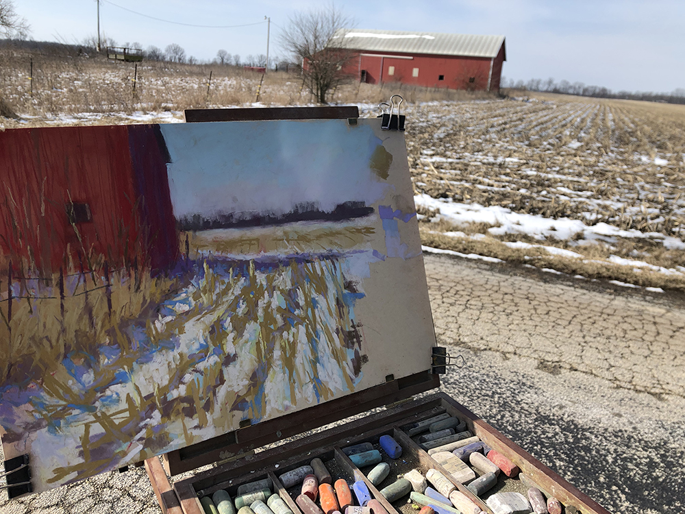 Small easel sitting outdoors holding a painting of a barn and field with the scene in the background