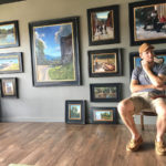 Artist sitting, posing with his wall of painitngs