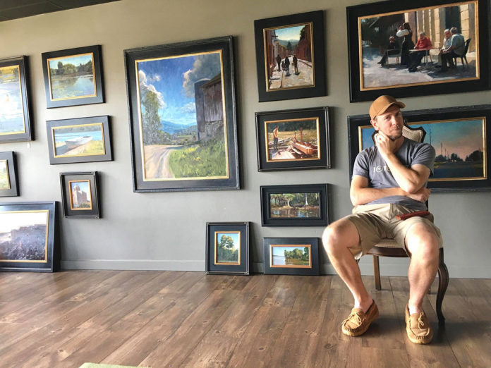Artist sitting, posing with his wall of painitngs