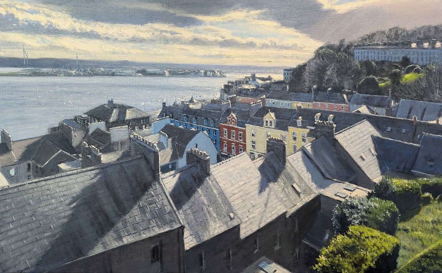 A view over rooftops of a coastal town, with rows of colorful houses, green hills, and sunlight reflecting on the water under a cloudy sky. 15th Annual PleinAir Salon Art Competition