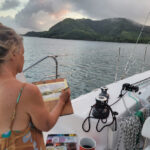 Forever Connected to a Moment Roxy Podlogar painting Malakati Village from the cockpit of her sailboat, 'Sky Pond'